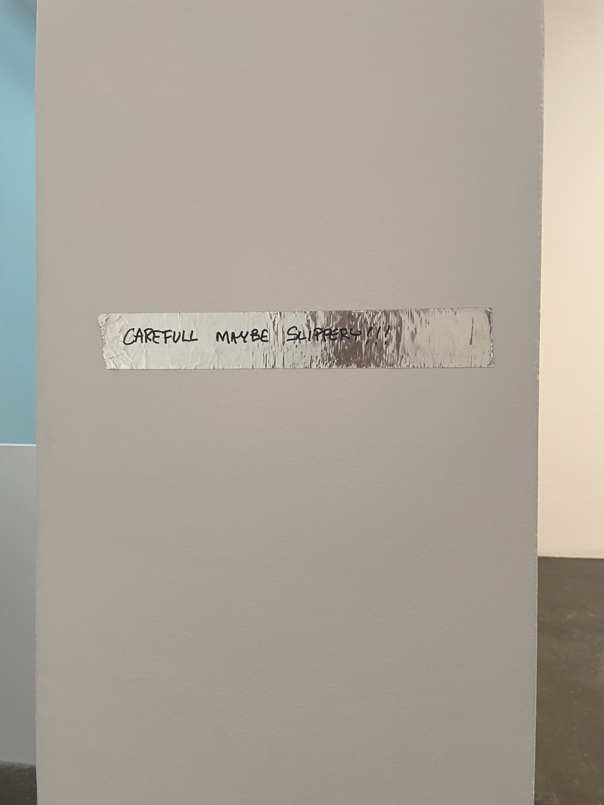 Devon Dikeou, Tape, from The Inconspicuouses at MCA Denver — a strip of silver tape on a gallery wall hand-lettered "CAREFULL MAYBE SLIPPERY!!!"